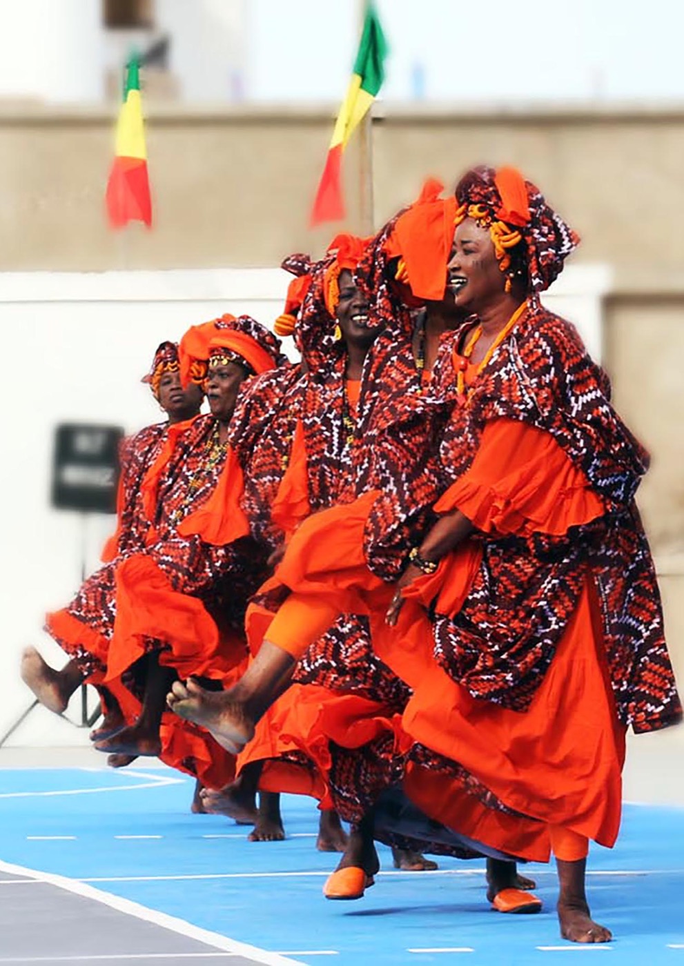 Bailarinas senegalesas con trajes y tocados naranjas y rojos en plena danza.